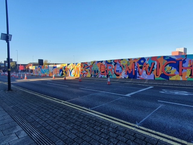 Colourful wall mural beneath blue sky in empty road, saying WEST BROMWICH