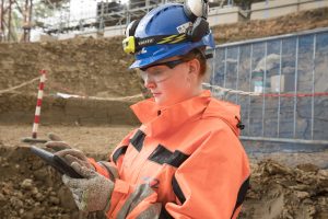 Young female engineer in a hard hat and reflective jacket on a construction site, looking at a tablet.