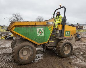 A woman in safety gear driving a large yellow JCB truck at a muddy construction site.