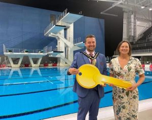The Mayor of Sandwell handing over a giant gold key to Joanna Rolfe of the Birmingham 2022 Commonwealth Games. They are standing in Sandwell Aquatics Centre next to a swimming pool.
